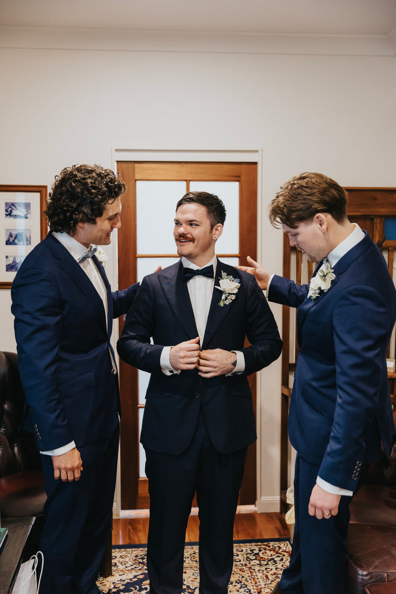 Three men in suits standing indoors, with one adjusting another's bow tie.